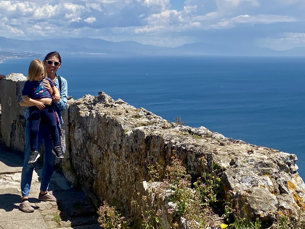 An adult and a child standing by a rock wall on the Rock of Gibraltar, with the African coastline visible across the Strait of Gibraltar.