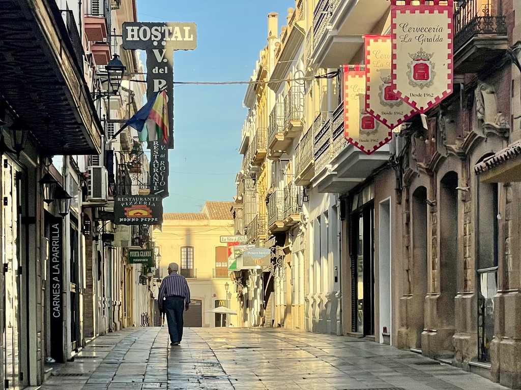 An empty cobblestone street in Ronda early in the morning, with shops and an elderly man walking.