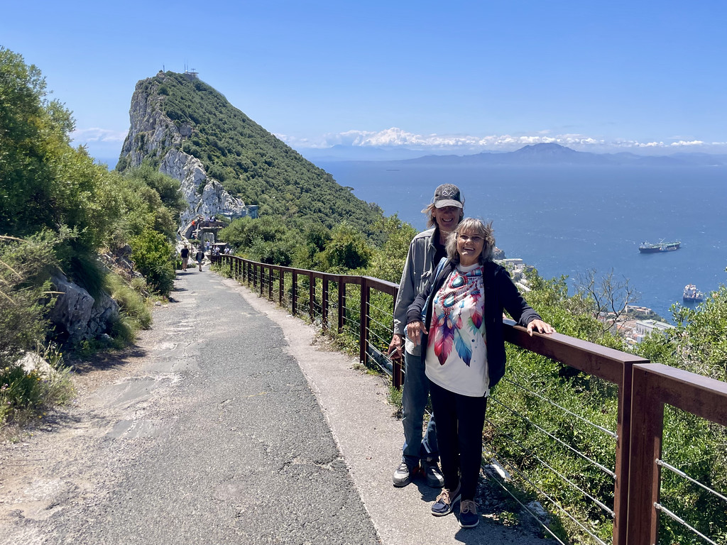 Two day trippers standing on a path on the Rock of Gibraltar, with the steep limestone peak behind them and views across the Strait of Gibraltar toward Africa.