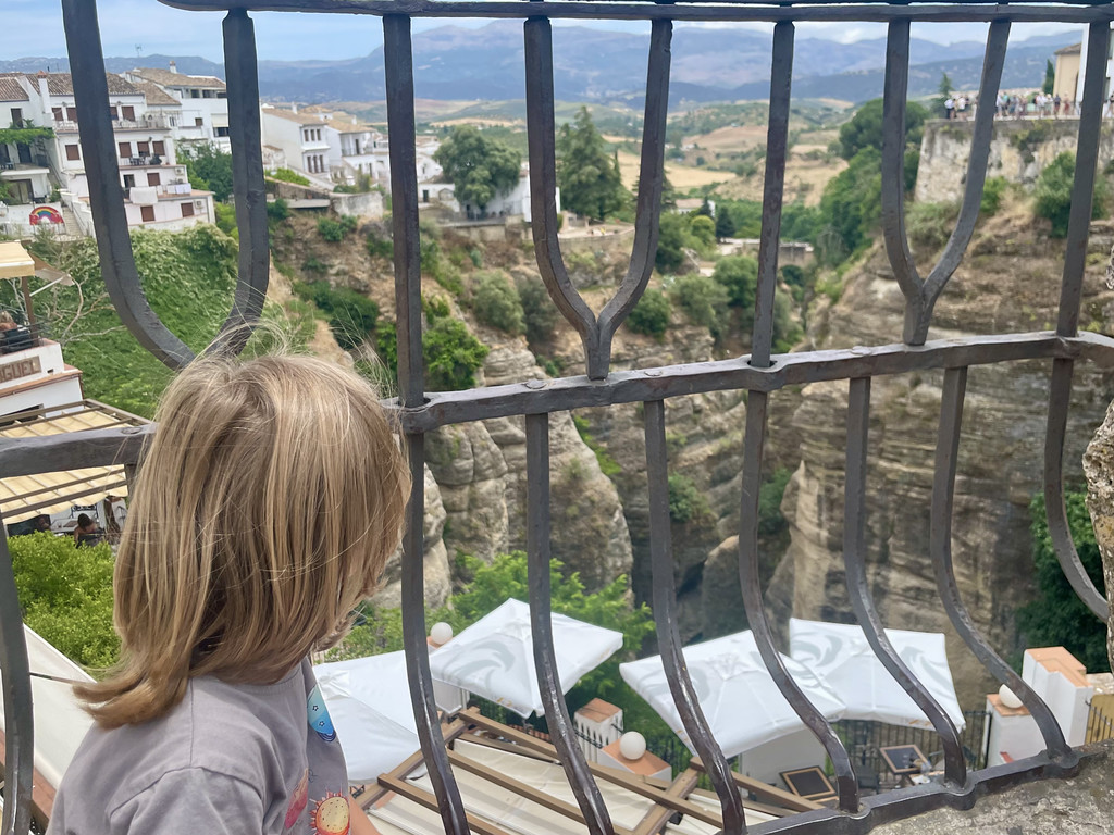 A child looking through a dark iron railing down at white buildings clustered on a cliffside.