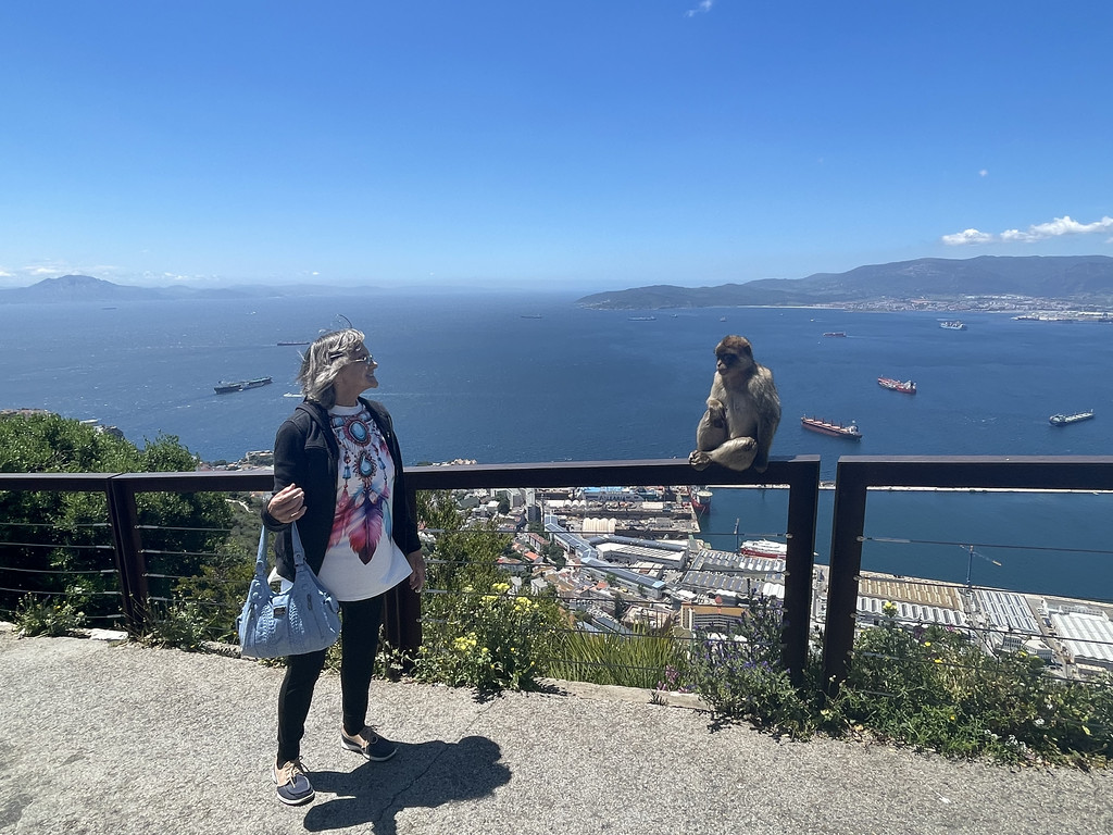 A day tripper standing near a Barbary macaque on a railing on the Rock of Gibraltar, with the Strait of Gibraltar, boats in the harbor, and the African coastline in the background.