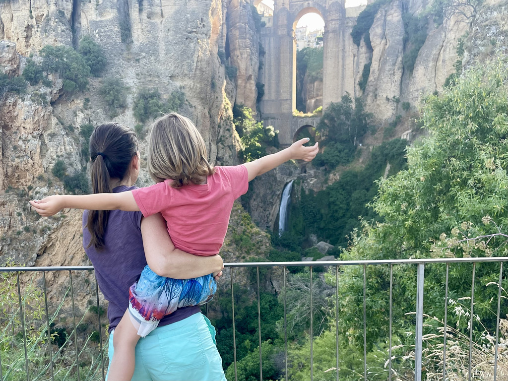 A parent holding a child, both pointing toward Puente Nuevo bridge and a waterfall, viewed from a lower gorge path.